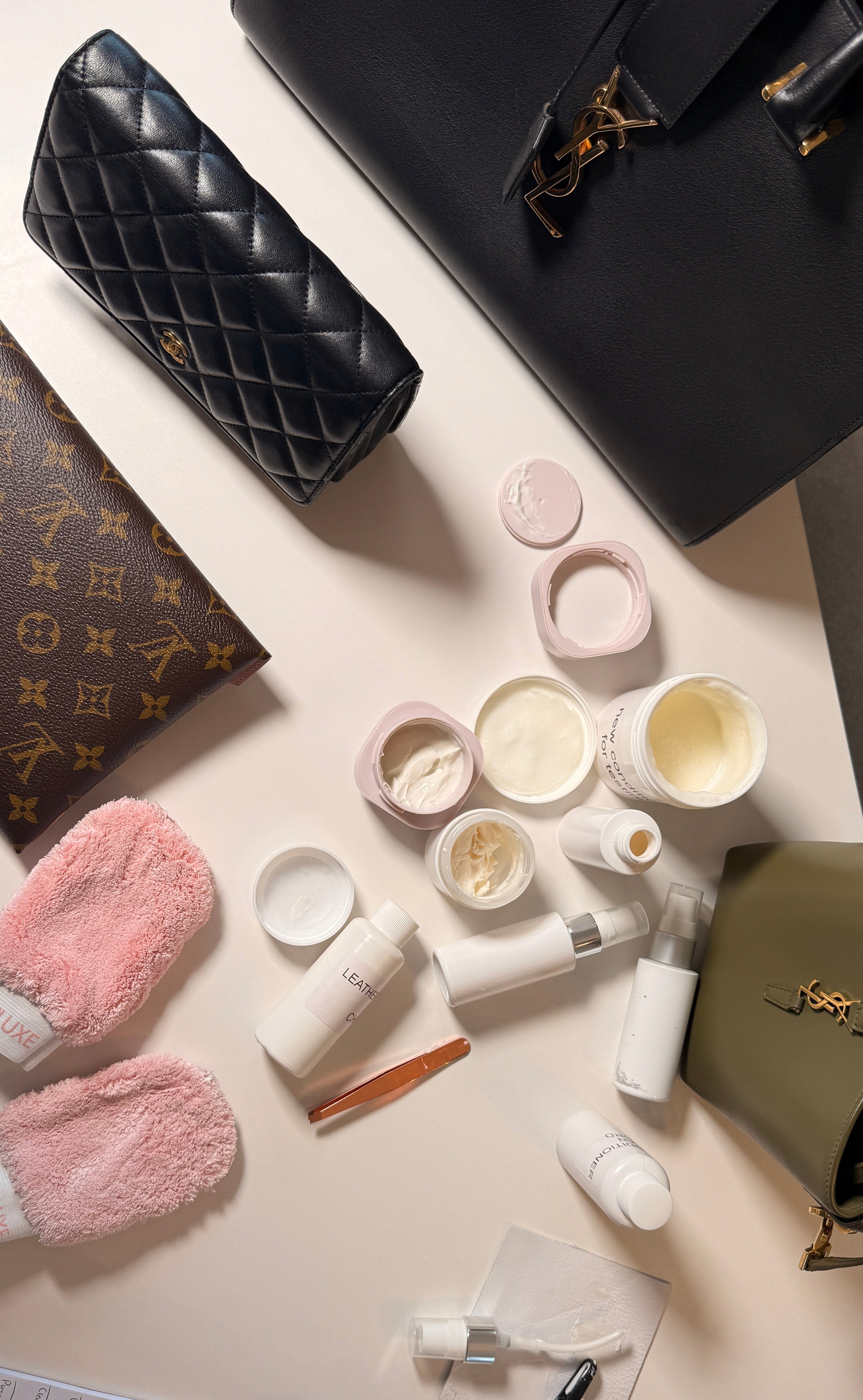 Flat lay of luxury handbags with skincare and cleaning products on a white surface, including pink slippers and accessories.